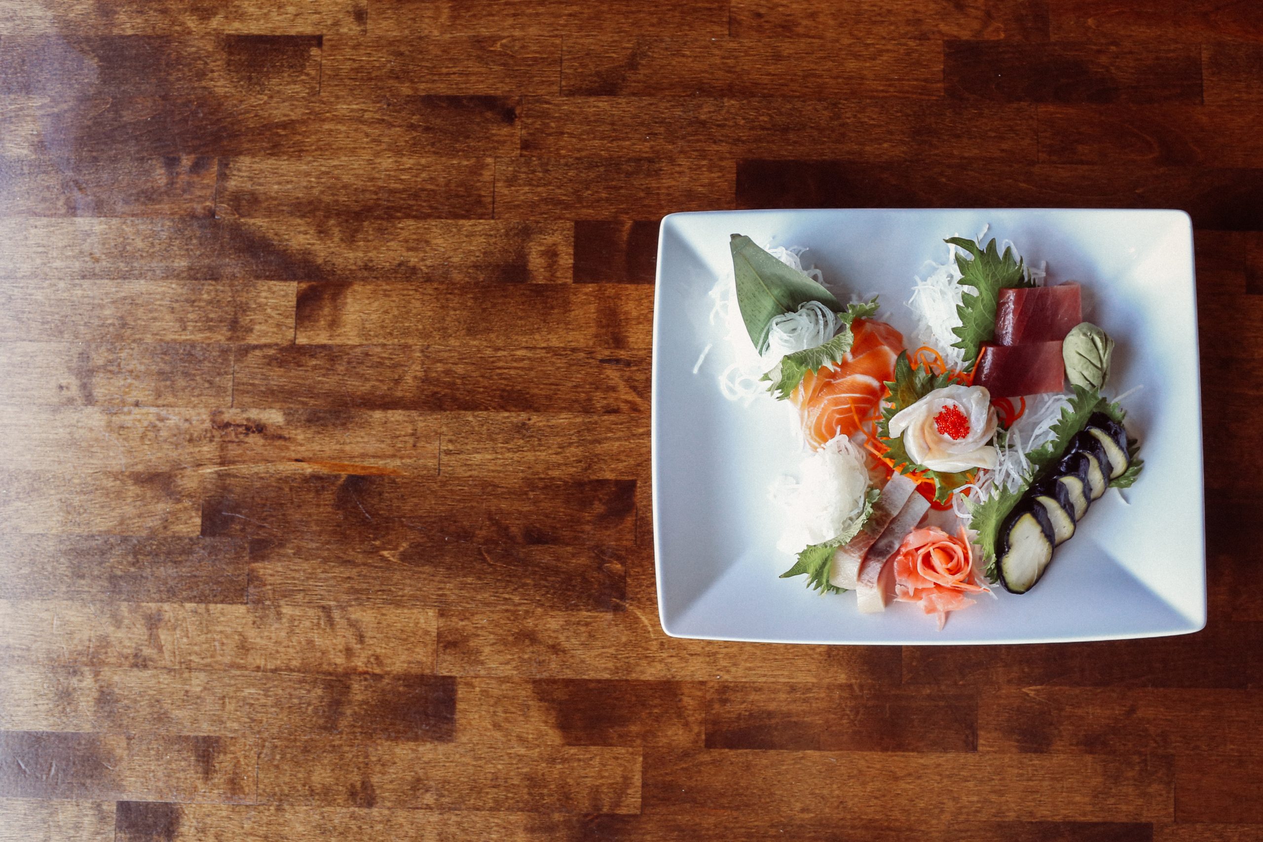 A beautifully-plated sushi entree sitting on the right hand of a wooden table.